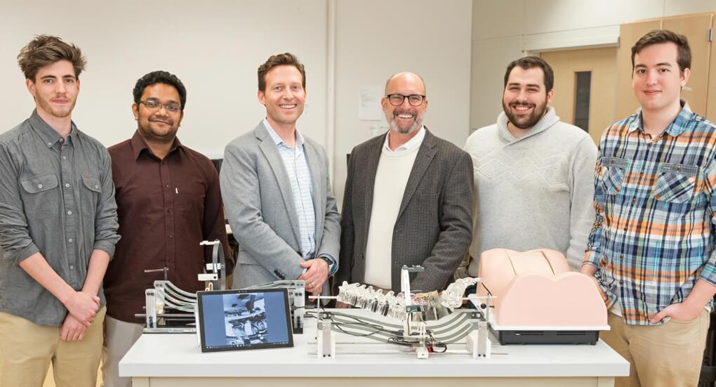 Staff and students from the aMDI/Encoris team that worked on the S2T Surgical Smart Trainer pose by the device.